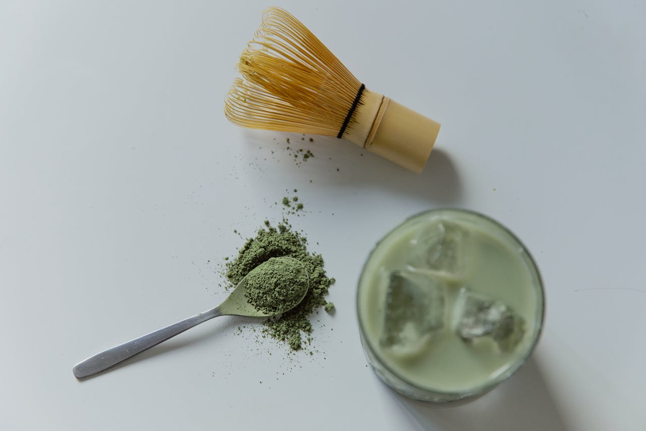 Top view of matcha latte, bamboo whisk, and powdered matcha on a white surface.